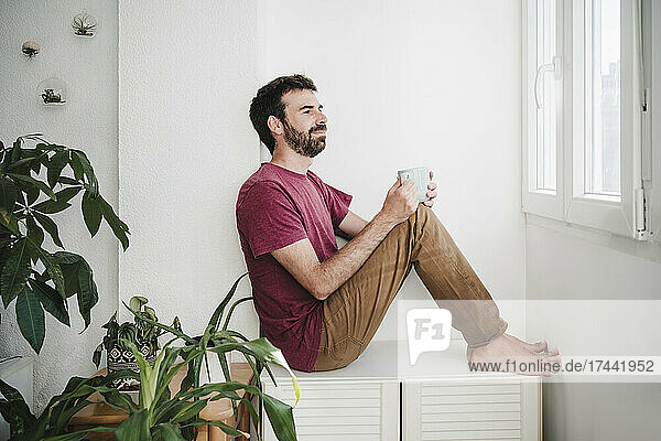 Thoughtful man with mug sitting on seat at home