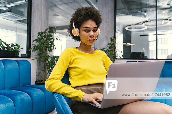 Businesswoman wearing wireless headphones while using laptop in office lobby