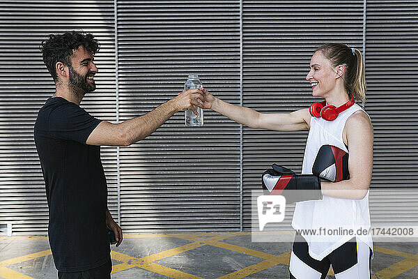 Smiling female boxer sharing water with male coach in front of wall