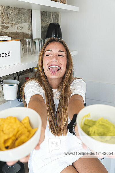 Glückliche Frau streckt die Zunge heraus  während sie in der Küche Nacho-Chips und Guacamole anbietet