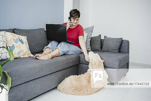 Businesswoman with laptop stroking dog while talking on smart phone at home