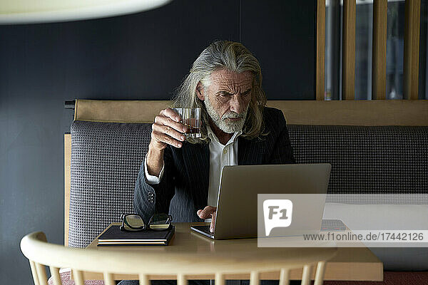 Male business person holding whiskey glass while working on laptop in hotel cafe