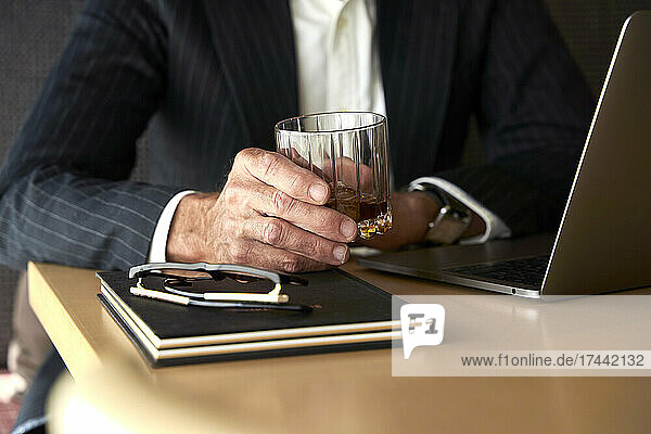 Mature businessman holding whiskey glass by laptop in hotel