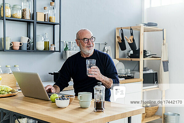 Älterer Mann mit Laptop hält Trinkglas zu Hause
