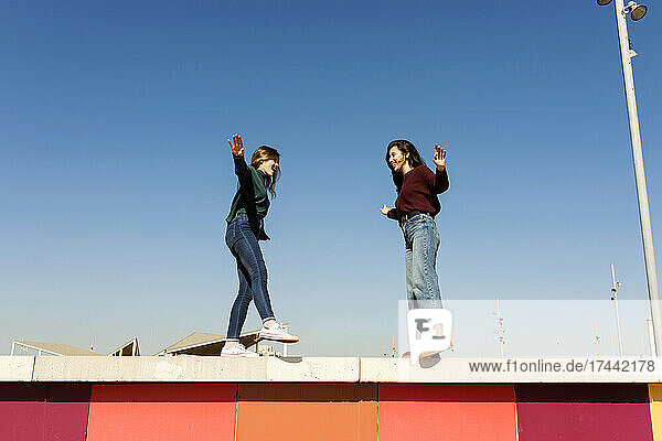 Playful female friends balancing while standing on retaining wall