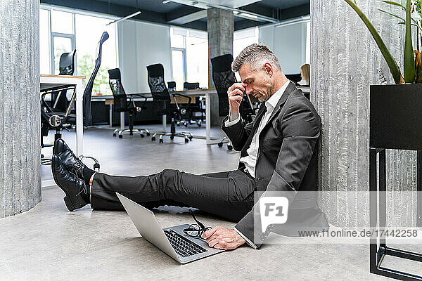 Tired businessman with laptop sitting on floor in office