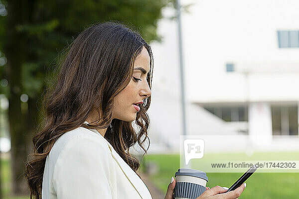 Woman holding disposable cup using mobile phone at park