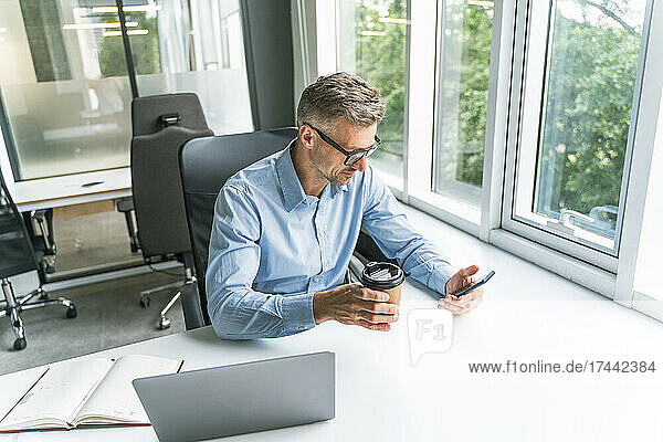 Businessman with coffee cup using mobile phone at desk in office