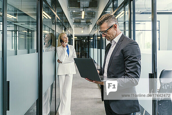 Geschäftsmann benutzt Laptop  während Kollegin im Flur im Büro mit dem Smartphone spricht