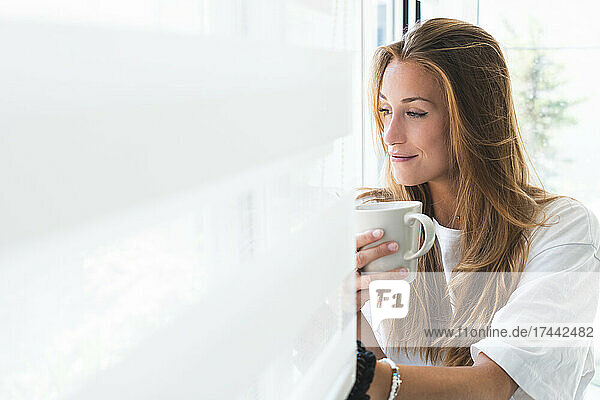 Schöne junge Frau mit Tasse  die zu Hause durch das Fenster schaut