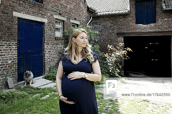 Pregnant woman touching abdomen while standing with dog in background at farmhouse