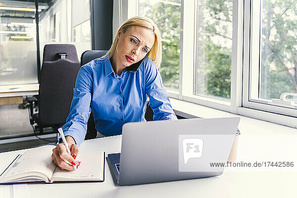 Blond businesswoman using laptop while talking on smart phone in office