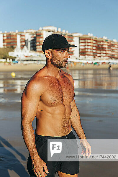 Mid adult shirtless man wearing cap standing at beach