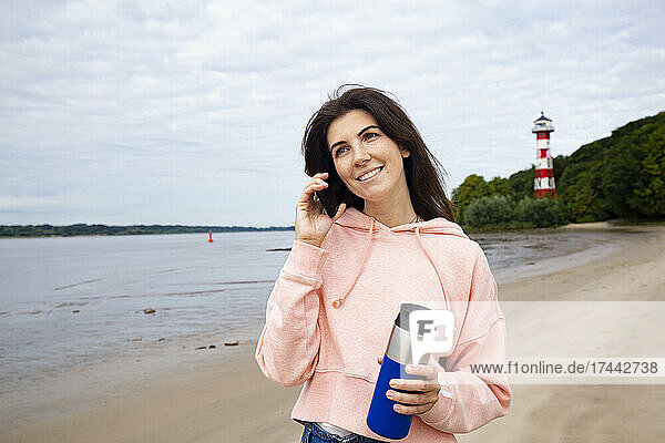 Lächelnde Frau  die am Strand mit einem Smartphone spricht und dabei einen isolierten Getränkebehälter in der Hand hält