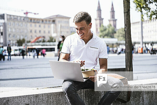 Smiling man with laptop having food in city