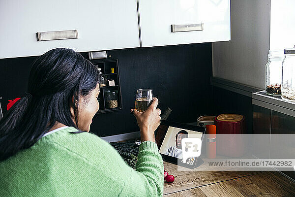 Frau mittleren Alters trinkt während eines Videoanrufs in der heimischen Küche