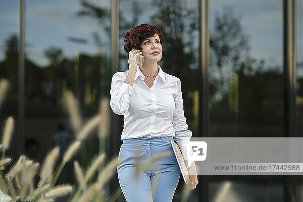 Businesswoman holding laptop while talking on smart phone outside office building