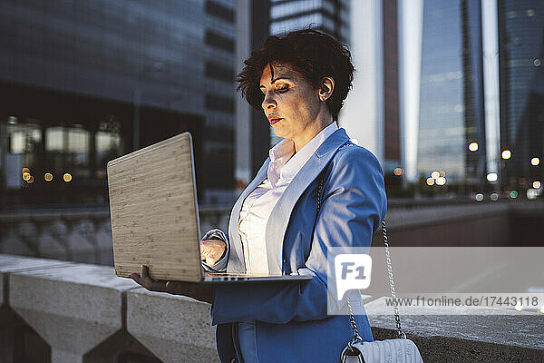 Berufstätige Frau benutzt Laptop  während sie in der Abenddämmerung am Geländer der Brücke steht