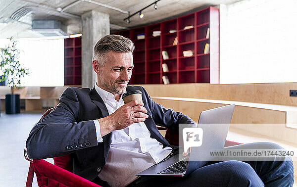Businessman holding disposable cup while using laptop in office