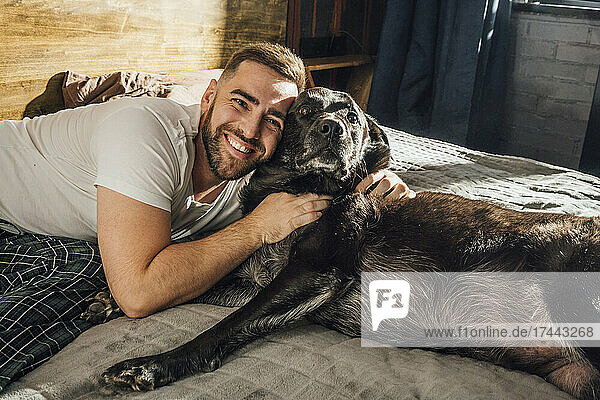 Lächelnder  gutaussehender Mann umarmt Hund zu Hause im Bett