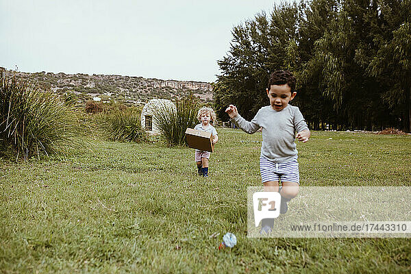 Boy running with male friend carrying cardboard box while playing on meadow