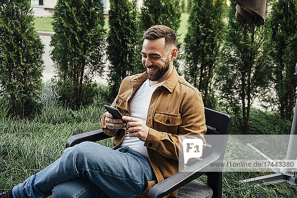 Lächelnder Geschäftsmann  der im Hinterhof Textnachrichten über sein Mobiltelefon sendet