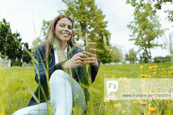 Smiling young businesswoman with mobile phone sitting in park