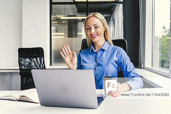 Smiling blond businesswoman waving hand during video call through laptop in office