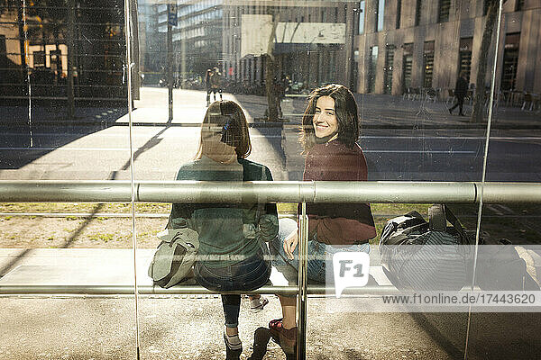 Lächelnde Frau blickt durch Glas über die Schulter  während sie mit ihrer Freundin an der Straßenbahnhaltestelle sitzt