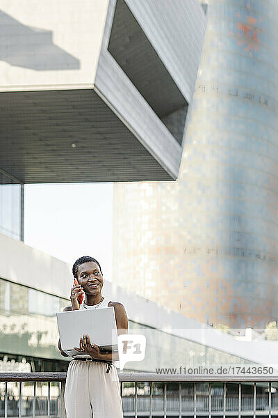 Lächelnde Geschäftsfrau mit Laptop  die am Geländer mit dem Mobiltelefon telefoniert
