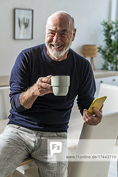 Cheerful man holding coffee cup and smart phone at home