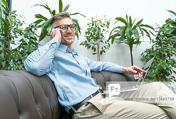 Smiling businessman with laptop talking on smart phone in office