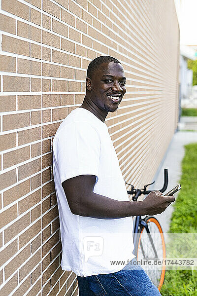 Smiling mid adult man with smart phone standing by brick wall