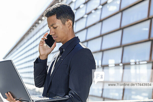 Businessman with laptop talking on smart phone outside office building during sunny day