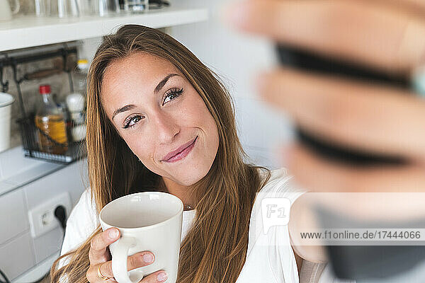 Beautiful woman taking selfie while holding mug at home