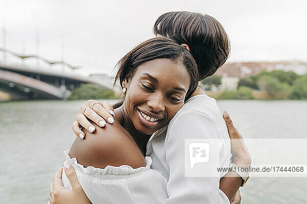 Smiling woman embracing girlfriend at lakeshore