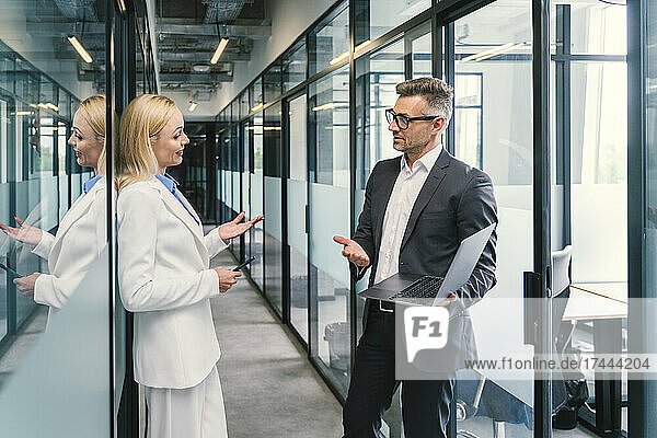 Businesswoman discussing with male colleague holding laptop in corridor at office