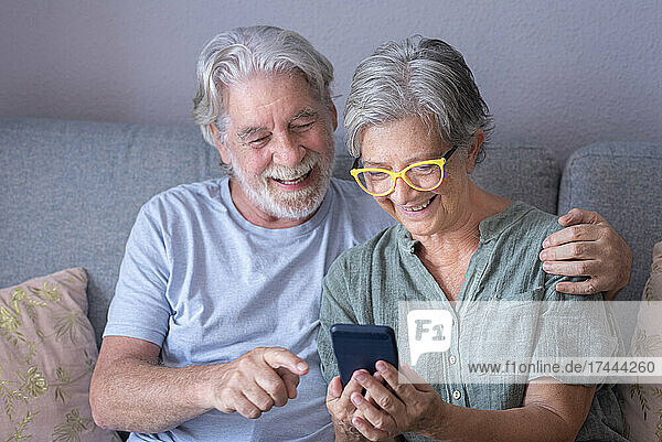Lächelnder bärtiger Mann zeigt auf sein Mobiltelefon  während er neben einer Frau im Wohnzimmer sitzt