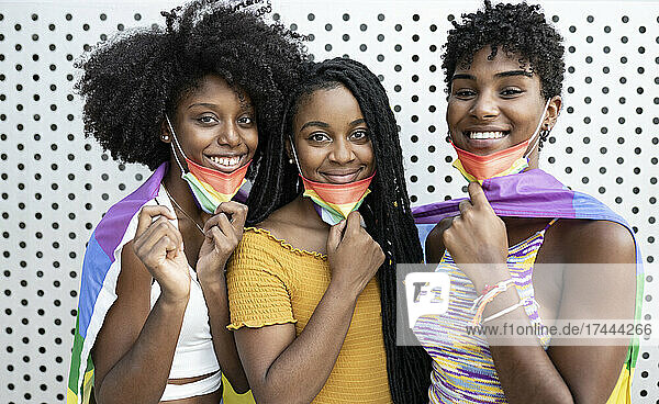 Lächelnde lesbische Frauen tragen schützende Gesichtsmasken mit Regenbogenflagge