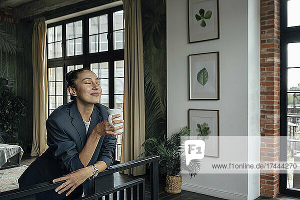 Female professional with eyes holding coffee cup while leaning on railing in office