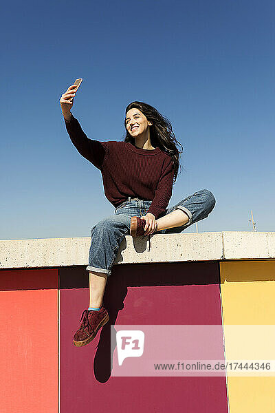 Young woman taking selfie through smart phone on retaining wall