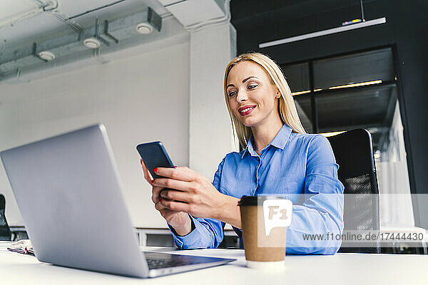 Geschäftsfrau benutzt Mobiltelefon  während sie mit Laptop im Büro sitzt