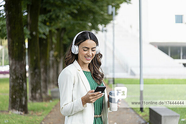 Glückliche Frau mit kabellosen Kopfhörern  die im Park ihr Mobiltelefon benutzt