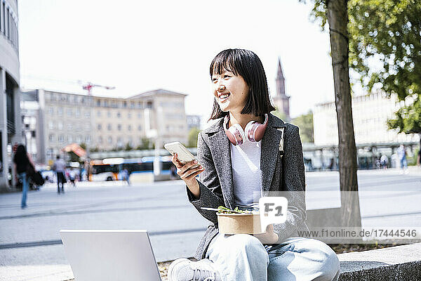 Smiling businesswoman with mobile phone and food sitting in city