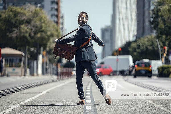 Happy businessman with crossbody bag spinning on road