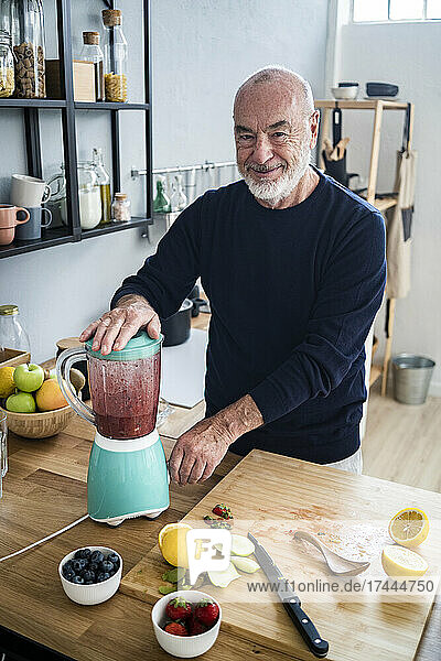 Smiling man preparing smoothie in electric juicer at kitchen counter