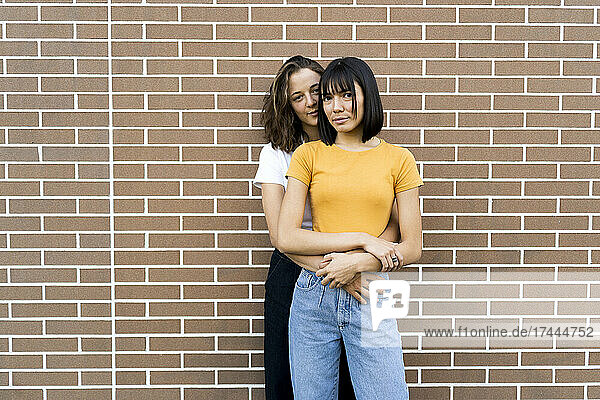 Young woman embracing girlfriend while standing in front of brick wall