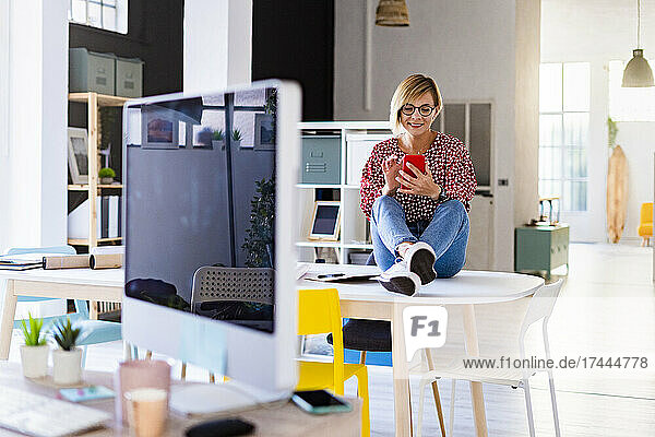 Lächelnde Berufstätige  die ihr Mobiltelefon benutzt  während sie am Schreibtisch sitzt
