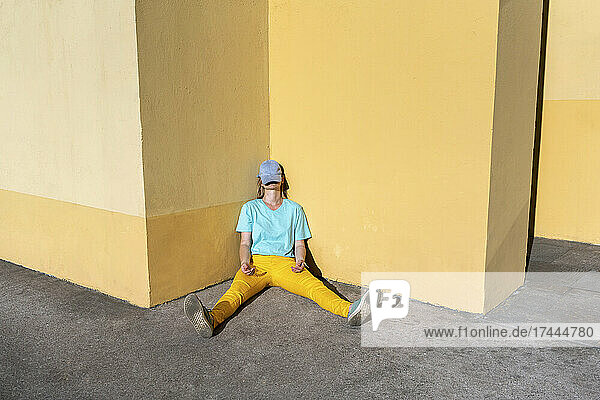 Tired mid adult woman sitting with cap on face by yellow wall