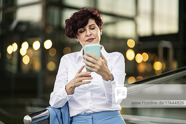 Mature businesswoman text messaging through mobile phone while leaning on railing